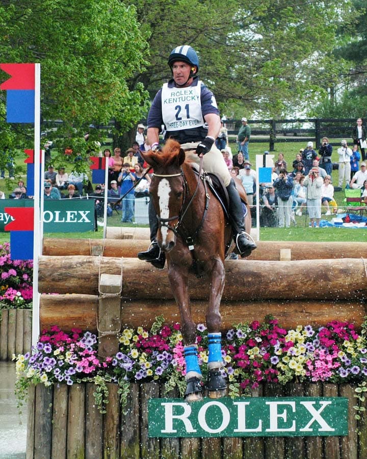 David O'Connor riding over a log jump into water on a cross country course while competing at the Rolex Kentucky Three-Day Event.