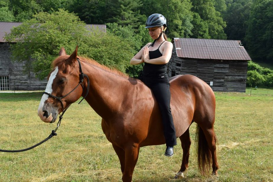 Cathy Woods practicing gratitude in prayer pose on a chestnut horse in a field with historic farm buildings in the background.